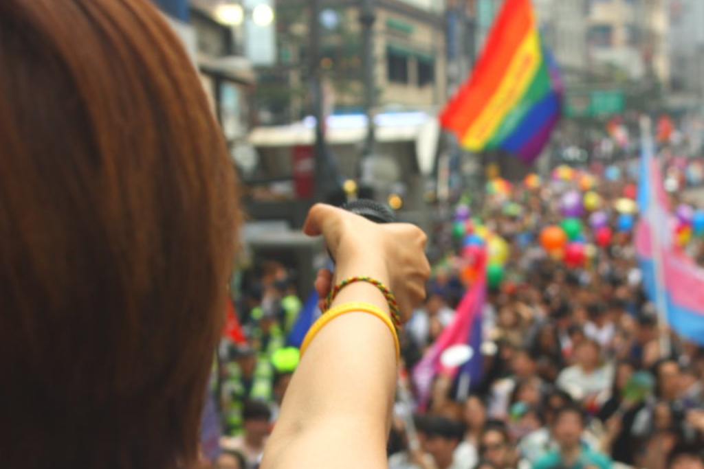 Behind right shoulder of someone holding microphone out over Pride Parade crowd LGBTQ+ equality
