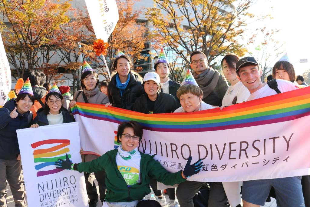 group of people pose together holding NIJIIRO DIVERSITY banners and wearing rainbow party hats LGBTQ+ equality