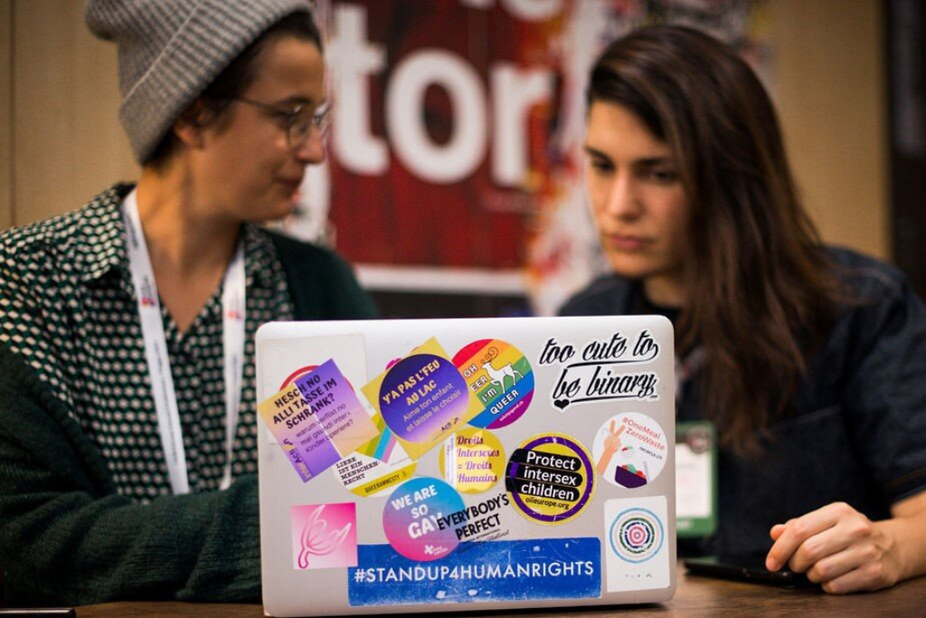 Two people sit behind a computer, the computer cover is covered with lgbtq+ pride stickers