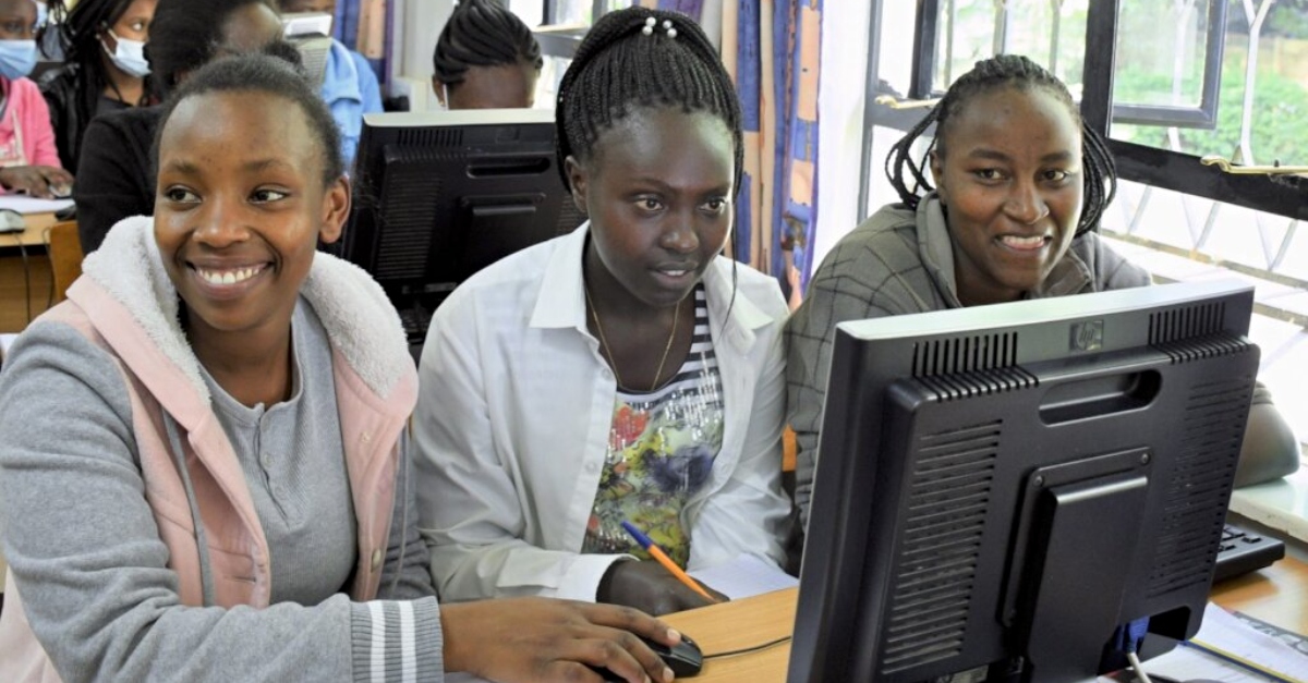 Three people sit at a desk behind a computer monitor.