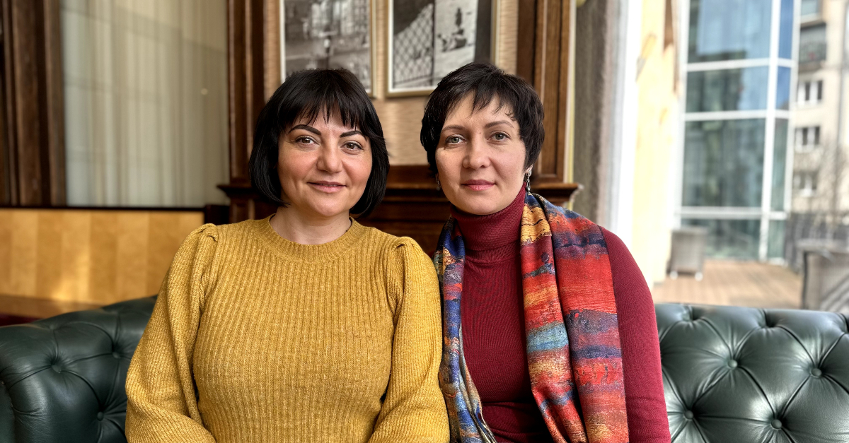 two women sit on a couch together, smile at the camera