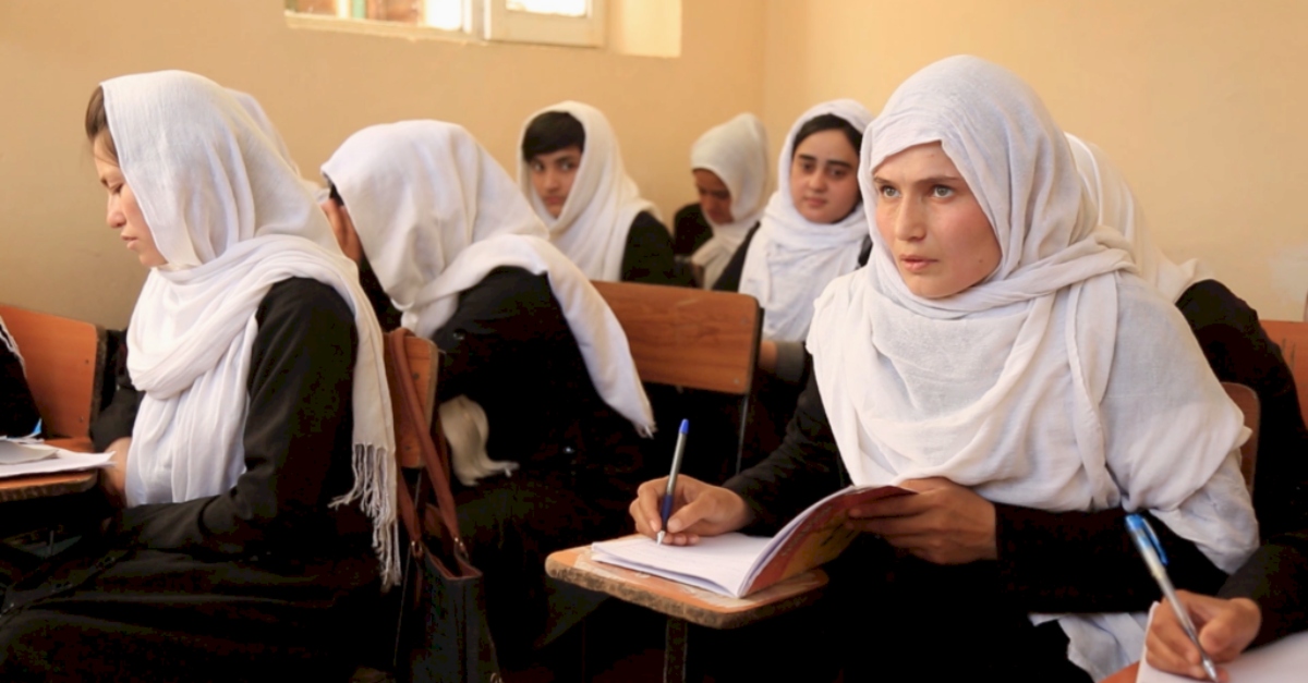 A student wearing a white hijab writes in a notebook at a wooden desk. Classmates wearing white hijabs are seated at desks around the student.