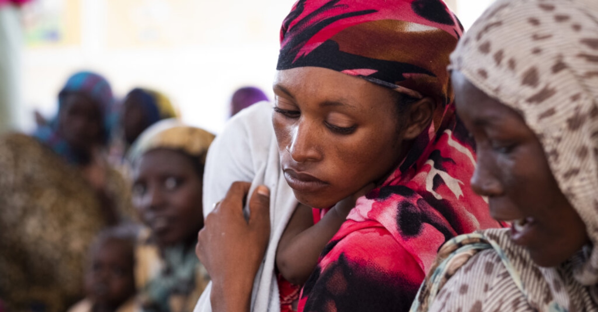 A person wearing a pink and black head covering holds a baby wrapped in a blanket over their shoulder during the Sudan conflict. Another person's face is out of focus in the foreground.