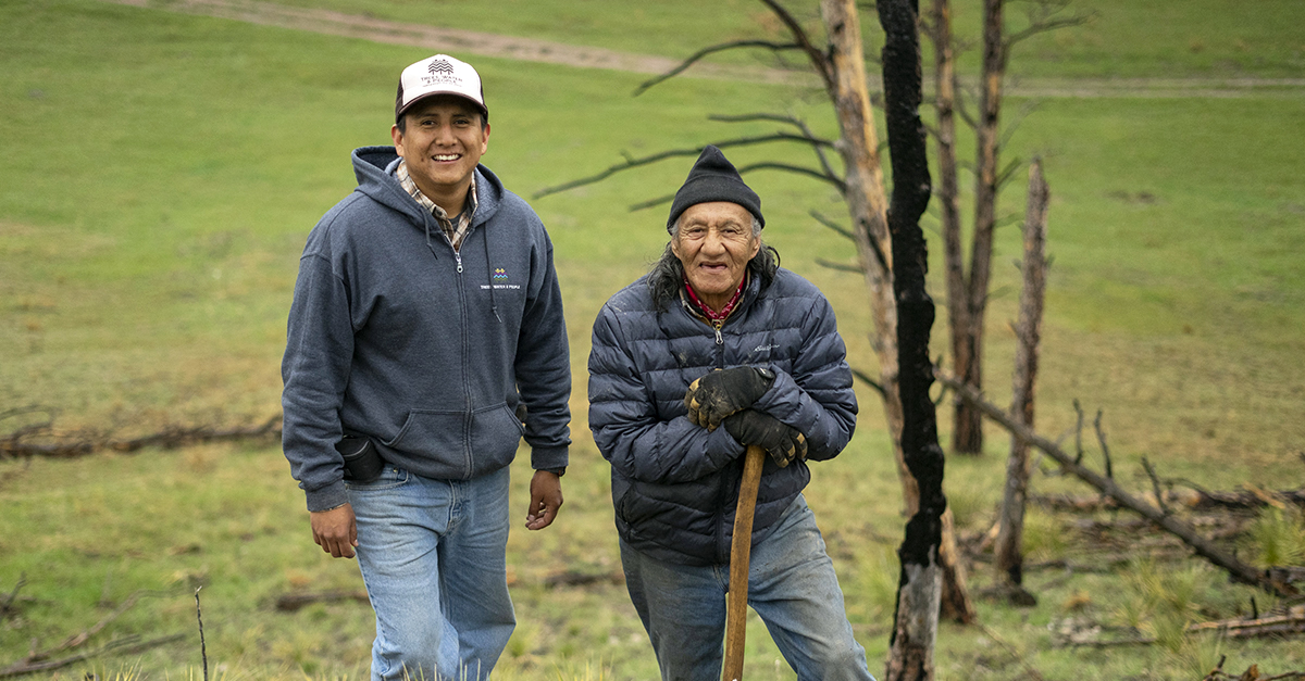 Two people wearing jackets and hats stand amid green grass and a few trees. The person on the right leans on the handle of a garden tool.