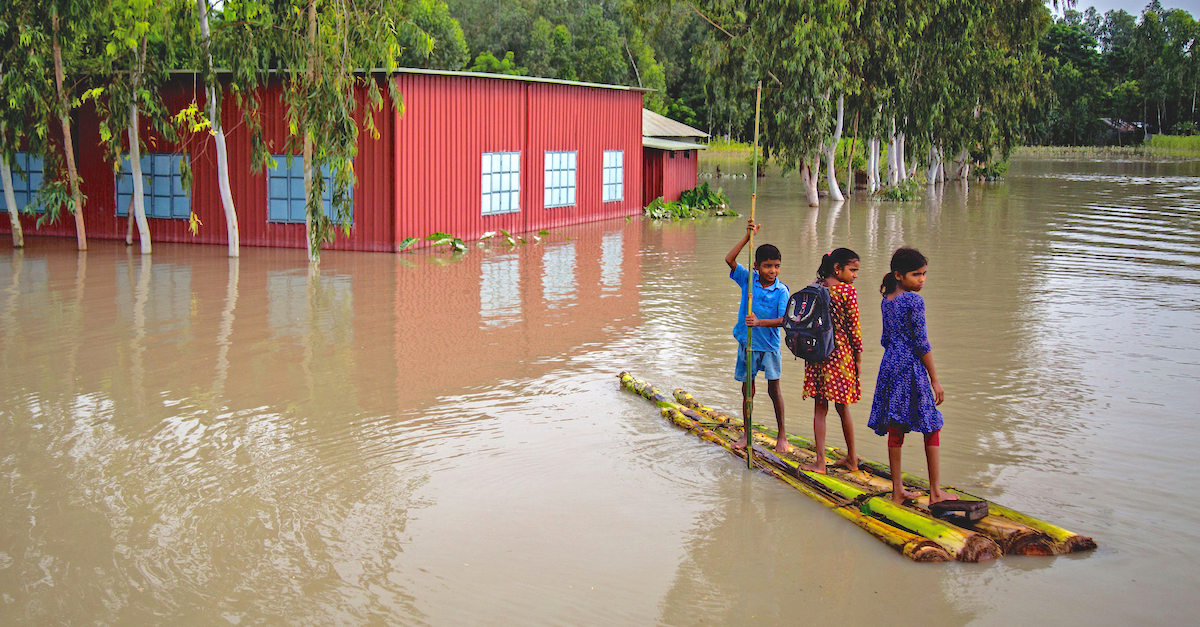 three kids, wearing school bags, navigate a flooded neighborhood on makeshift raft