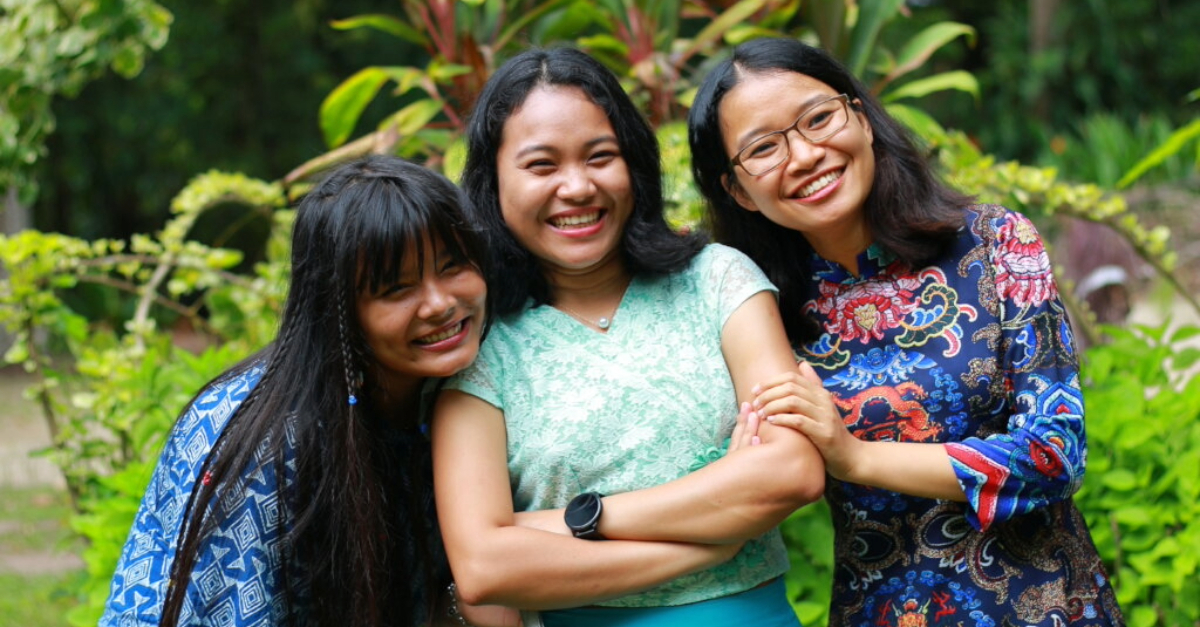 Three women smiling at the camera in a green setting with trees and plants.