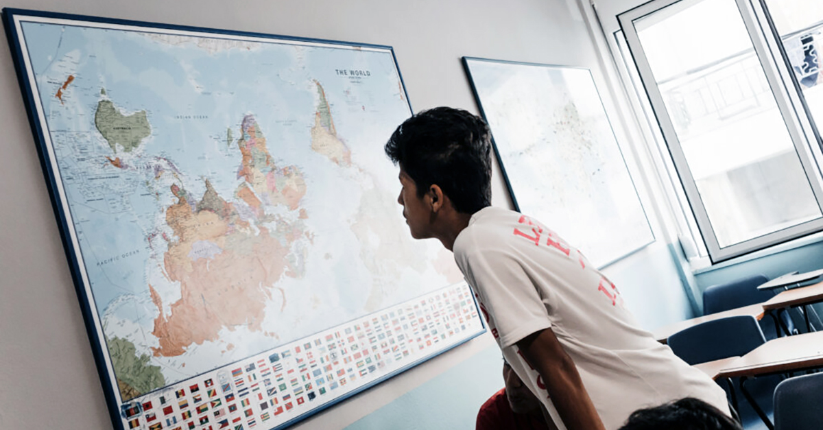 A young man in a white T-shirt looks at a map of the world. Photo by Better Days Greece.