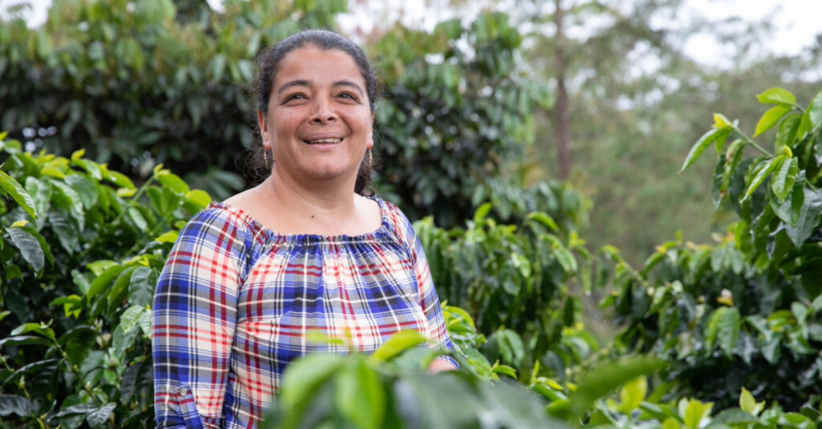 A woman smiling stands slightly left of centre surrounded by green foliage.