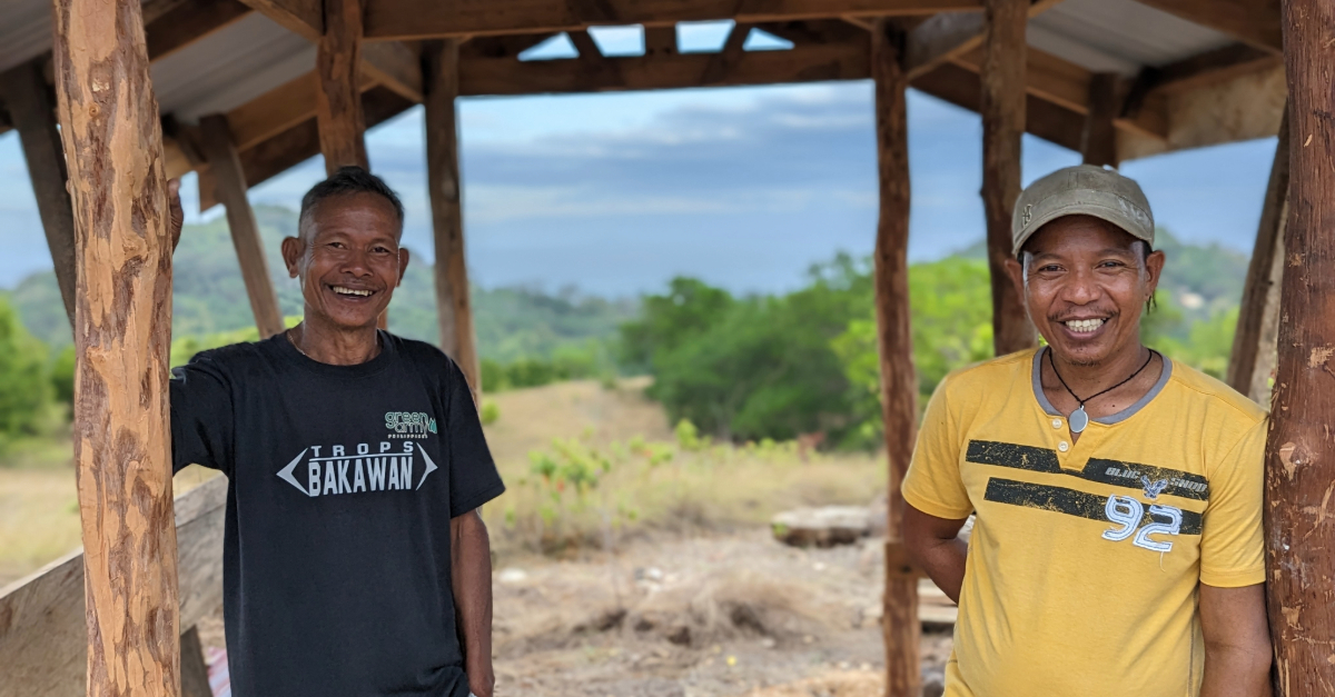 Two men are stood inside a wooden structure, on either side of the frame smiling and facing the camera. They have beautiful scenery of trees and hills behind them.