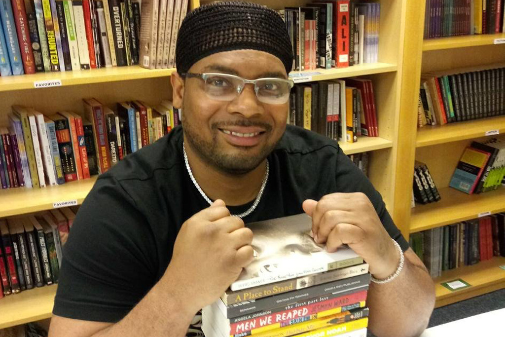 A man sitting down at a table, smiling with his hands on a stack of books, with a bookcase in the background.
