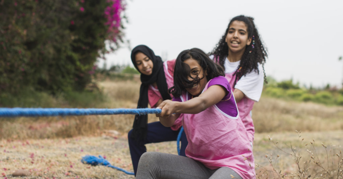 Three girls in pink jerseys pull a blue tug-of-war rope with smiles on their faces