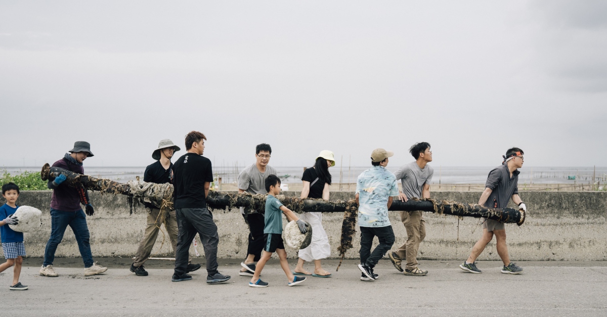 This image shows a group of people working together to carry a long, debris-covered pipe along a coastal road.