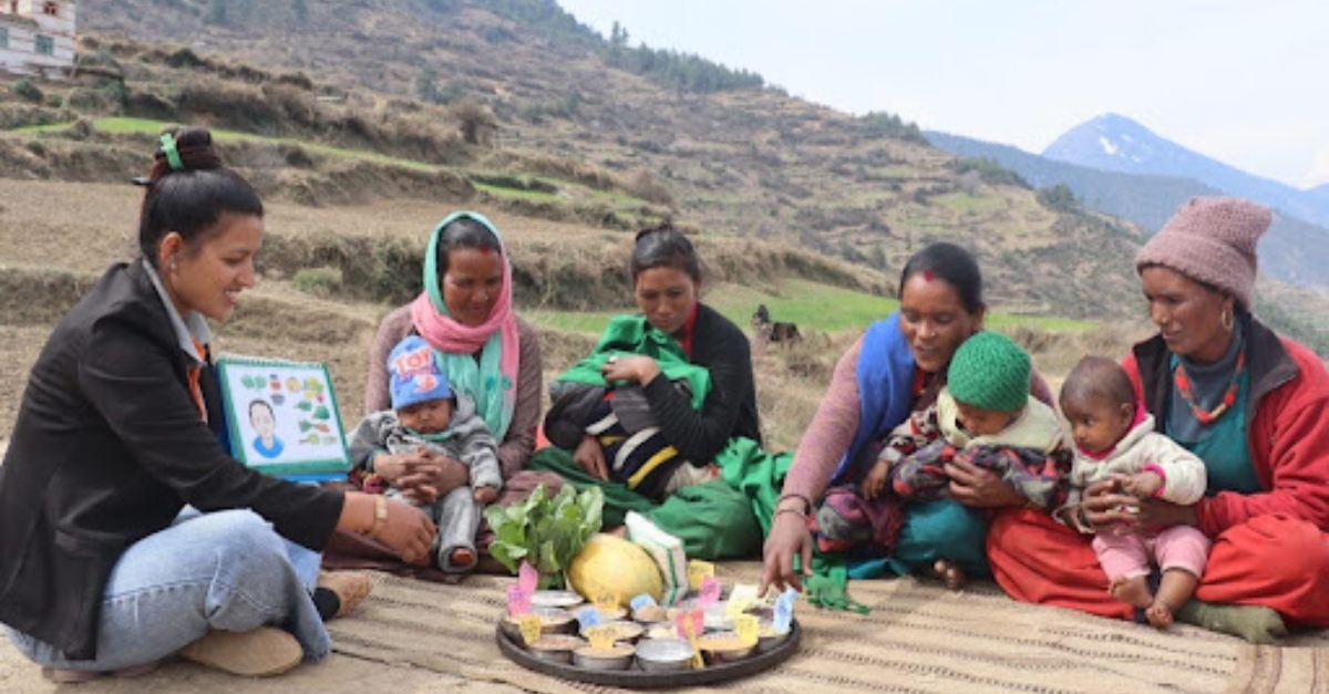 A group of five women and three young children sit together on a woven mat in a rural, mountainous setting, gathered around a tray of food and vegetables. One woman holds up an illustrated nutrition chart.