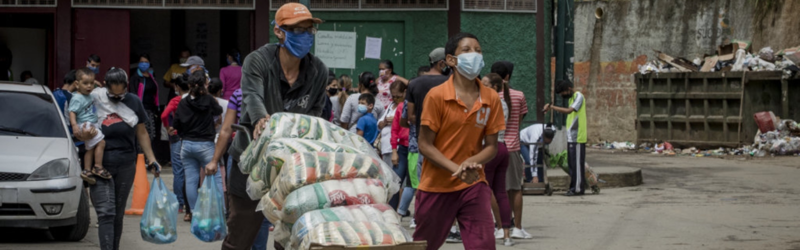 A man pushes a cart with food supplies as a boy walks next to him. Families walk behind holding plastic shopping bags.